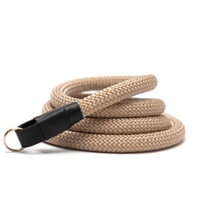 Peut inclure: Une corde beige enroulée avec un connecteur en cuir noir et un anneau en métal argenté. La corde est épaisse et semble être faite de fibres tissées. Le fond est blanc.