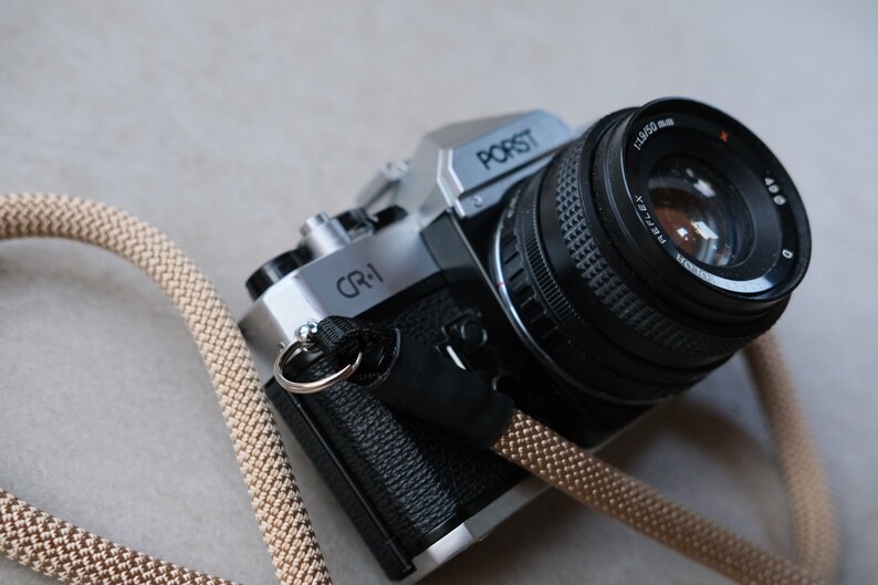May include: A silver and black Porst CR-1 35mm film camera with a black lens and a tan camera strap. The lens is marked with "1:1.9/50 mm" and "Reflex". The camera is on a neutral background.