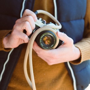 May include: A classic black and silver camera with a beige strap is held. The lens reflects a warm, colourful scene. The person is wearing a navy blue vest with a white zip and a mustard-coloured sweater. The camera is a CR-1 model.