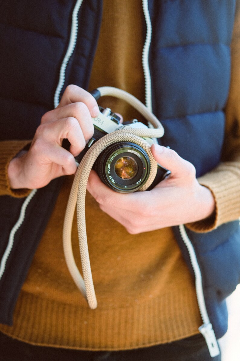 May include: A classic black and silver camera with a beige strap is held. The lens reflects a warm, colourful scene. The person is wearing a navy blue vest with a white zip and a mustard-coloured sweater. The camera is a CR-1 model.