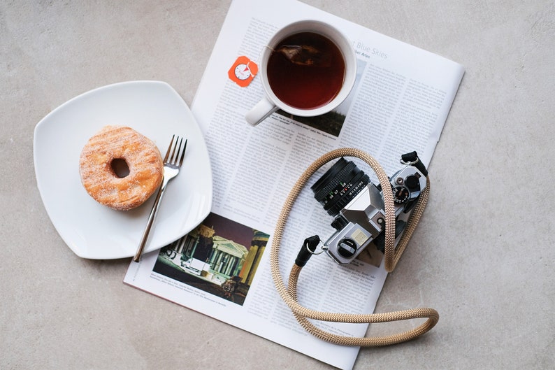 Könnte beinhalten: Draufsicht einer Frühstücksszene mit einem gezuckerten Donut auf einem weißen Teller mit einer Gabel, einer Tasse Tee und einer Vintage-Kamera mit einem beigefarbenen Riemen auf einer Zeitung.