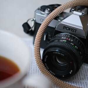May include: A close-up of two vintage cameras, one silver and black, the other black. A tan camera strap is looped around the lens of the front camera. The lens is black with white text. A cup of tea is in the foreground.