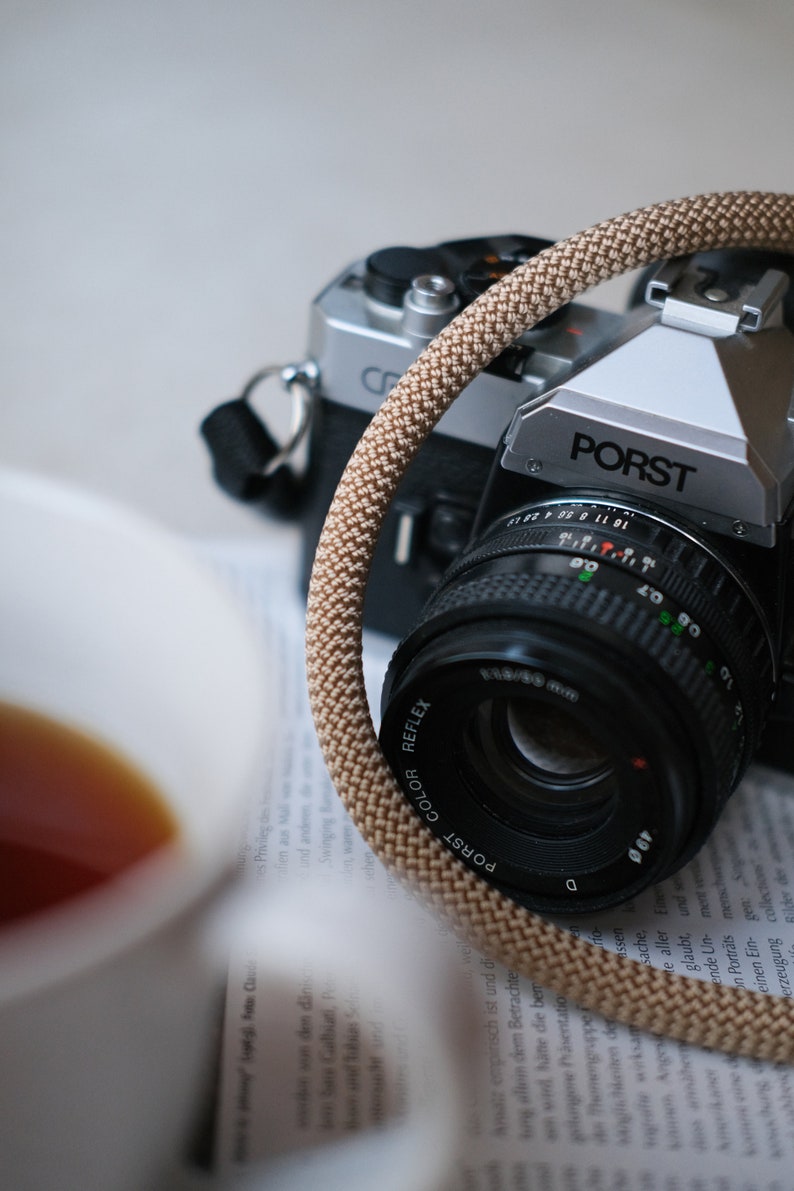 May include: A close-up of two vintage cameras, one silver and black, the other black. A tan camera strap is looped around the lens of the front camera. The lens is black with white text. A cup of tea is in the foreground.