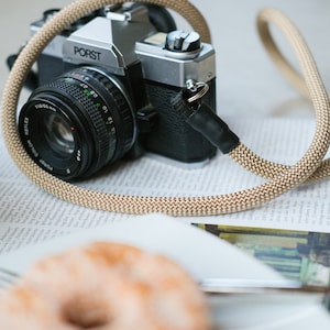 May include: A classic PORST camera with a beige rope strap sits on a newspaper. The camera features a black lens and silver details. A donut is in the foreground, hinting at a lifestyle or food photography theme.