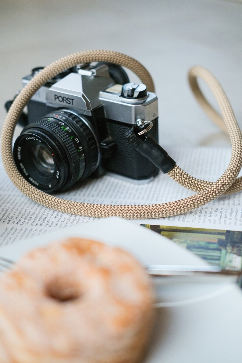 May include: A classic PORST camera with a beige rope strap sits on a newspaper. The camera features a black lens and silver details. A donut is in the foreground, hinting at a lifestyle or food photography theme.