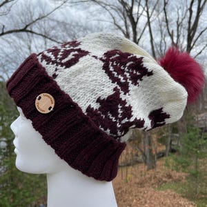 May include: A knit beanie featuring a cream and maroon geometric pattern with a ribbed maroon cuff. A round wooden tag with the text Loonstruct Creations is attached to the cuff, and a fluffy maroon pom-pom sits at the top of the hat.