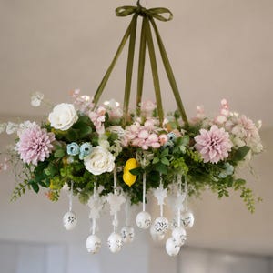 May include: A hanging floral chandelier featuring a circular arrangement of pink dahlias, white roses, green foliage, and a single yellow lemon. White eggs with black patterns hang from the base on ribbons, suspended by olive green ribbons.