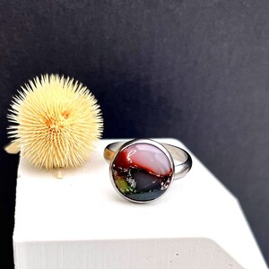 Könnte beinhalten: Ein silberner Ring mit einem runden, mehrfarbigen Stein, der in der Mitte eingesetzt ist. Der Stein hat Farbtöne von Rot, Orange, Gelb, Grün und Schwarz. Der Ring wird auf einer weißen Plattform präsentiert.