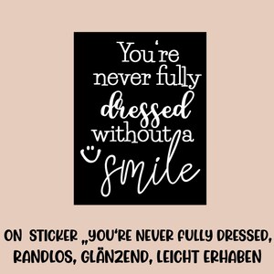 Könnte beinhalten: Ein schwarzes Quadrat mit dem weißen Text "You're never fully dressed without a smile." Der Hintergrund ist hellpfirsichfarben. Der Aufkleber trägt auch den Text "RUB - ON STICKER".
