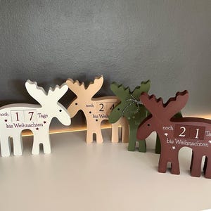 Könnte beinhalten: Eine Sammlung von Elch-förmigen Weihnachtskalendern aus Holz in Weiß, Beige, Grün und Braun. Jeder Elch zeigt eine Zahl an, die die Tage bis Weihnachten angibt, mit Text auf Deutsch. Die Kalender sind mit Sternen verziert.
