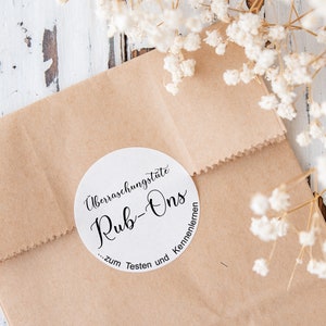 Könnte beinhalten: Eine braune Papiertüte mit einem weißen runden Aufkleber, auf dem "Überraschungstüte Rub-Ons ... zum Testen und Kennenlernen" steht.