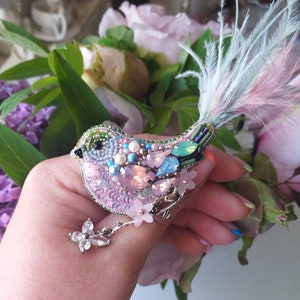 Könnte beinhalten: Handgefertigte Vogelbrosche mit einem rosa paillettenbesetzten Körper, verziert mit blauen und rosa Kristallen, Perlen und Federn. Der Vogel hat einen silbernen Zweig mit kleinen rosa Blüten und einem Kristallschmetterling. Die Brosche wird in einer Hand gehalten.