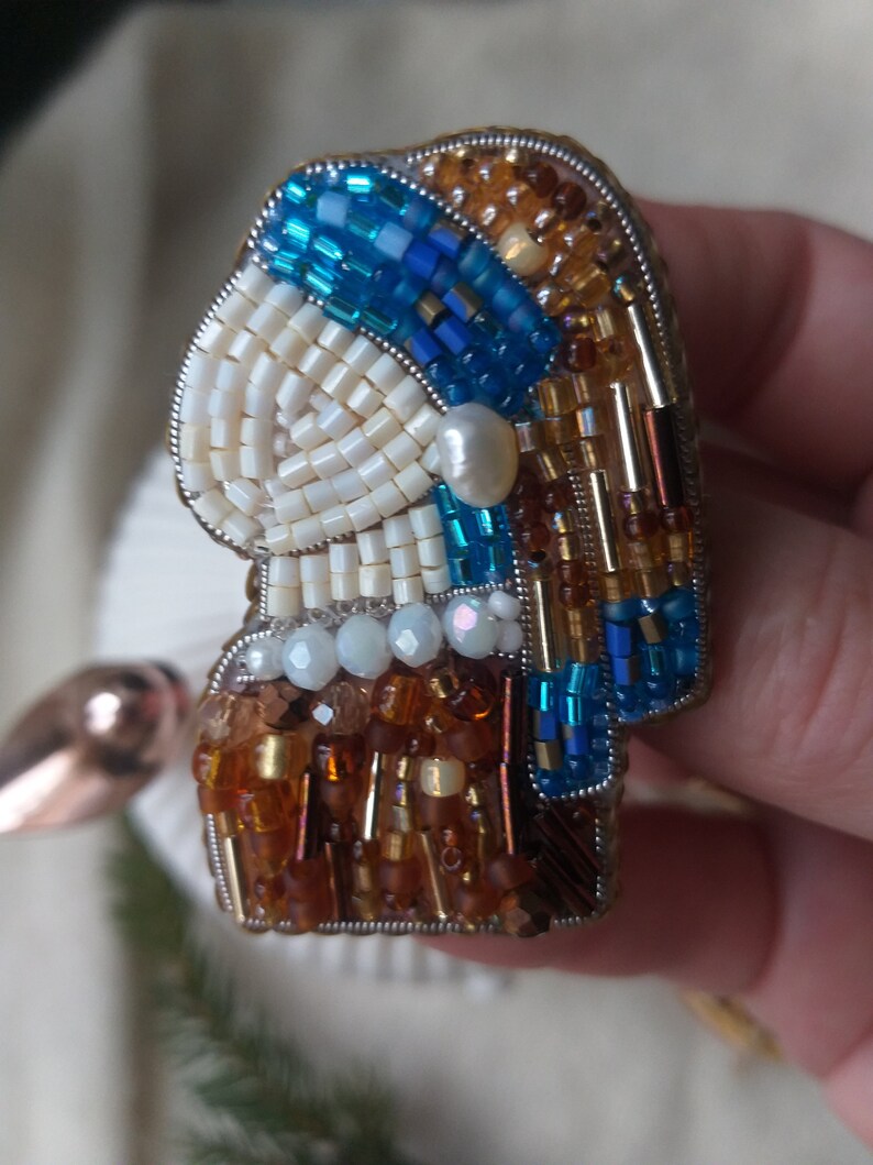 K&ouml;nnte beinhalten: Eine Perlenbrosche mit einem Frauenportr&auml;t, bestehend aus blauen, wei&szlig;en und braunen Perlen. Die Brosche ist aus verschiedenen Perlen gefertigt, darunter Rocailles, Bugle-Perlen und Perlen.