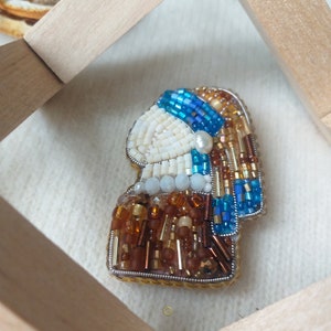 K&ouml;nnte beinhalten: Eine Perlenbrosche, die ein Portr&auml;t einer Frau mit einem blauen und goldenen Turban darstellt. Die Brosche ist aus wei&szlig;en, blauen, goldenen und braunen Perlen gefertigt.