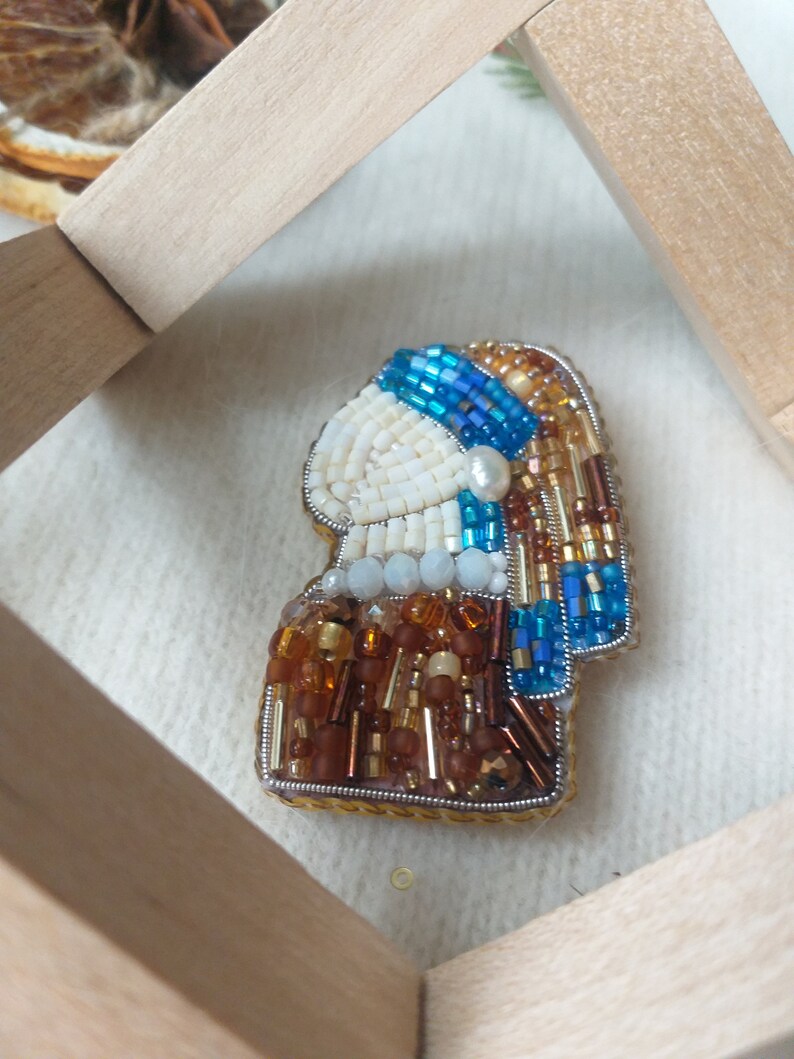 K&ouml;nnte beinhalten: Eine Perlenbrosche, die ein Portr&auml;t einer Frau mit einem blauen und goldenen Turban darstellt. Die Brosche ist aus wei&szlig;en, blauen, goldenen und braunen Perlen gefertigt.