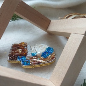 K&ouml;nnte beinhalten: Eine Brosche in Form eines Stiefels mit braunen, wei&szlig;en, blauen und goldenen Perlen. Die Brosche wird in einem Holzrahmen pr&auml;sentiert.