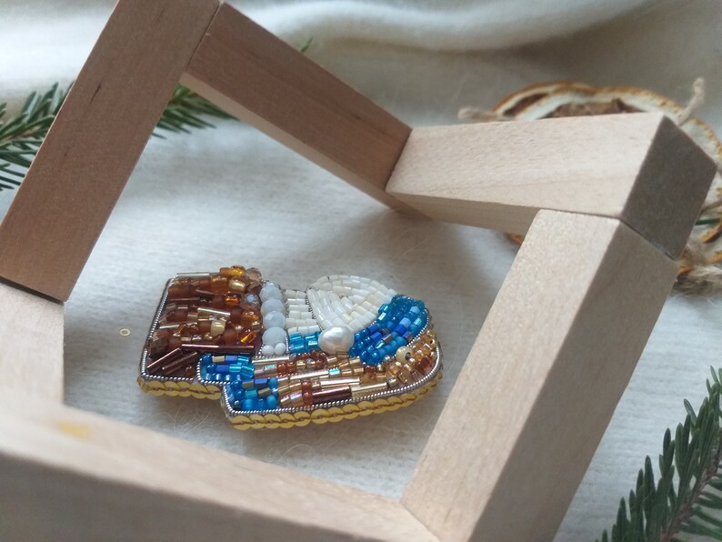 K&ouml;nnte beinhalten: Eine Brosche in Form eines Stiefels mit braunen, wei&szlig;en, blauen und goldenen Perlen. Die Brosche wird in einem Holzrahmen pr&auml;sentiert.