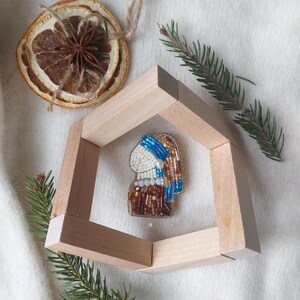 K&ouml;nnte beinhalten: Eine Perlenbrosche in Form eines Frauenkopfes mit einem blau-wei&szlig; gemusterten Kopftuch. Die Brosche wird in einem Holzrahmen pr&auml;sentiert.