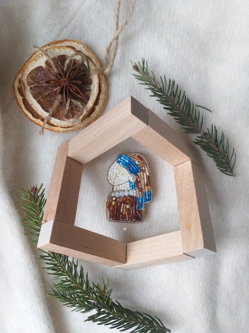 K&ouml;nnte beinhalten: Eine Perlenbrosche in Form eines Frauenkopfes mit einem blau-wei&szlig; gemusterten Kopftuch. Die Brosche wird in einem Holzrahmen pr&auml;sentiert.