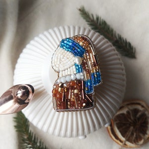 K&ouml;nnte beinhalten: Eine Perlenbrosche mit dem Portr&auml;t einer Frau mit einem blauen Kopftuch und einem Perlenohrring. Die Brosche ist aus wei&szlig;en, blauen, braunen und goldenen Perlen gefertigt.