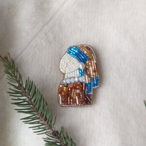 K&ouml;nnte beinhalten: Eine Perlenbrosche, die das Profil einer Frau mit einem blau-wei&szlig;en Kopftuch und einem Perlenohrring darstellt. Die Brosche ist aus braunen, wei&szlig;en und blauen Perlen gefertigt.