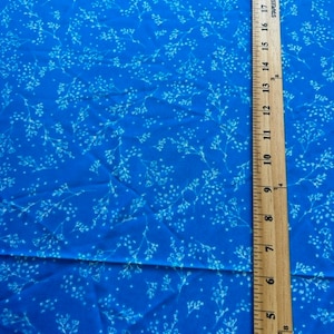 May include: Bright blue fabric with a delicate white floral pattern. A wooden ruler with inch measurements is placed vertically along the right side of the fabric, showing measurements up to 21 inches.