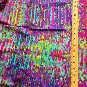 May include: Vibrant fabric with a colorful zigzag pattern in shades of purple, teal, pink, and yellow. A wooden ruler with inch markings is visible along the right side of the fabric, showing measurements.