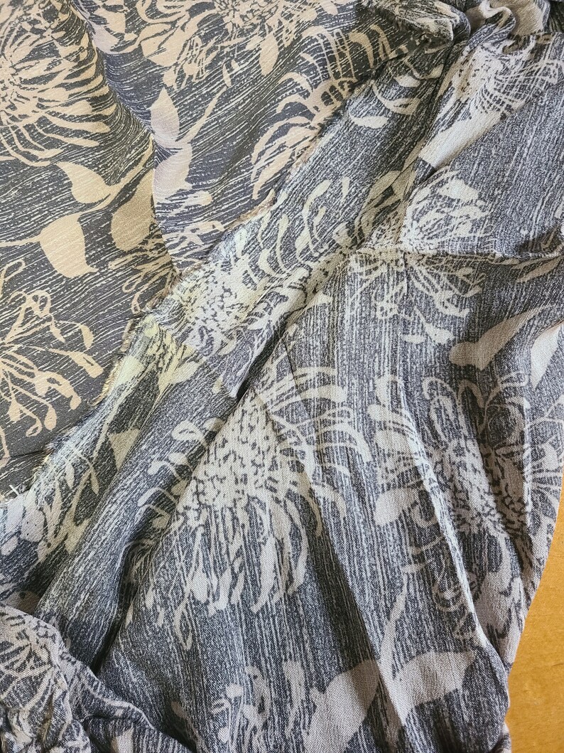 May include: A close-up of a gray and white patterned fabric with a floral design. The fabric is soft and flowing, and the pattern is subtle and elegant.
