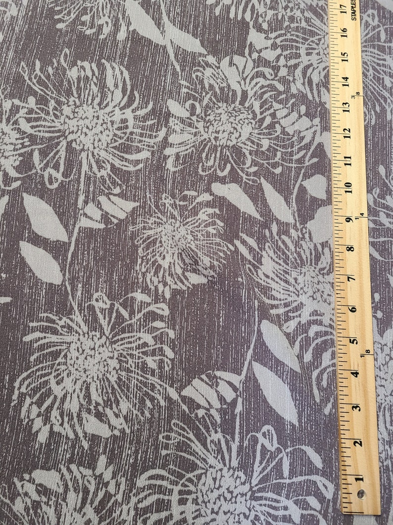 May include: A close-up of a brown fabric with a white floral pattern. The fabric has a textured, linen-like appearance. A ruler is visible in the bottom right corner of the image.