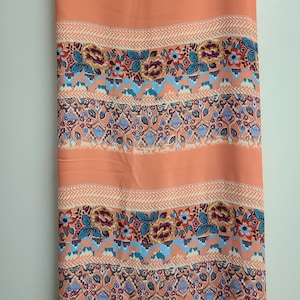 May include: Peach-colored fabric with a repeating pattern of floral and geometric designs. The fabric features horizontal stripes with alternating patterns of flowers, geometric shapes, and textured lines. The color palette includes peach, blue, purple, and cream.