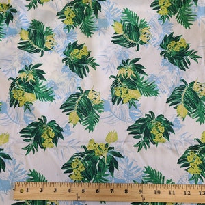 May include: White fabric with a repeating pattern of green and yellow floral clusters and green leaves. Light blue leaf silhouettes are also visible. A wooden ruler is placed across the bottom of the fabric, measuring up to 13 inches.