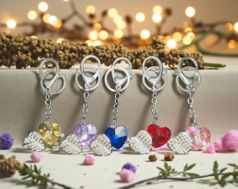 May include: Five keychains with heart-shaped pendants in yellow, purple, blue, red, and pink. Each heart is embellished with rhinestones and has silver-colored wings. The keychains have silver clasps and chains.