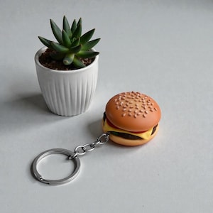 May include: A novelty burger keychain with a silver keyring. The miniature burger has a sesame seed bun, a tomato slice, cheese, and a burger patty. A small succulent plant in a white pot is in the background.