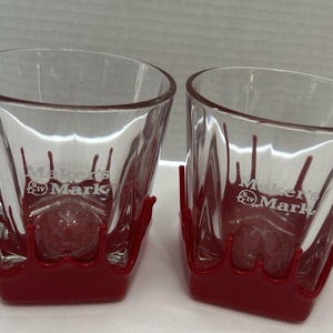 May include: Two clear, angled-sided drinking glasses with a red base. The glasses have the "Maker's Mark" logo printed on the front. The red base has a unique design that appears to be dripping down the sides of the glass.