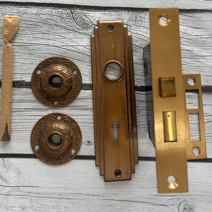 Può includere: Set di ferramenta per porte in bronzo antico, tra cui due placche rotonde a rosetta con motivi decorativi, una placca frontale rettangolare con un buco della serratura e un meccanismo di chiusura. È presente anche un piccolo oggetto avvolto.