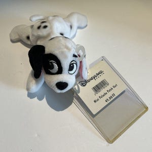 May include: A small plush Dalmatian dog with black spots, black ears, and a black patch over one eye. The toy is lying on a white surface with a Disneyland Paris tag attached.
