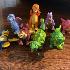 May include: A collection of colorful dinosaur figurines in various poses. The toys include a blue dinosaur with red wings, a standing orange dinosaur, and several green, purple, and pink dinosaurs. The toys are on a wooden surface.
