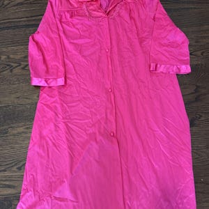 May include: A bright pink, knee-length robe with a button-down front and three-quarter sleeves. The robe features a collar with embroidered detailing and satin trim on the cuffs. The fabric appears lightweight and flowing.