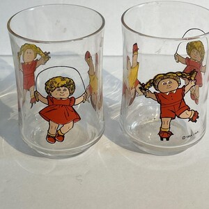 May include: Two clear drinking glasses featuring a cartoon girl in a red dress and shoes, jumping rope. The girl has blonde pigtails and is smiling. The glasses have a cylindrical shape and are set against a white background. The copyright date is 1984.