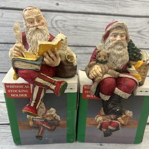 May include: Two whimsical Santa Claus stocking holders. One Santa is reading a book and holding a quill, the other is holding a teddy bear. Both are dressed in red and white suits and are sitting on boxes.