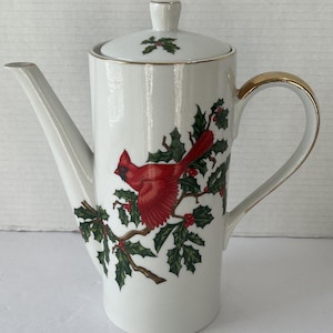 May include: White ceramic coffee pot with a lid, featuring a red cardinal bird perched on a holly branch with red berries. The handle and rim are trimmed with gold. A festive holiday-themed design.