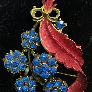 May include: Vintage floral brooch featuring clusters of blue rhinestones. The brooch has a red enamel leaf and a gold-tone bow. The design is reminiscent of a bouquet, with a classic, elegant style. The pin is a perfect accessory for a jacket or dress.
