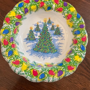 May include: A white ceramic Christmas serving bowl with a scalloped edge. The bowl features a festive design of Christmas trees and colorful lights. The rim is decorated with green garland and red, yellow, blue, and purple lights.