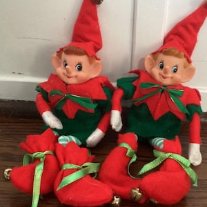 May include: Two elf dolls with peach-colored faces, pointed ears, and brown hair. They wear red hats with bells, green and red outfits with bows, white gloves, and red curled shoes with green ribbons and small bells attached to the tips.