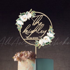 May include: A gold-colored cake topper with the names "The Keighley" and the date "10.26.2022" in a circular design, adorned with white flowers and green leaves. The topper is placed on a multi-tiered cake with floral decorations.