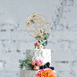 Peut inclure: Un cake topper en bois avec les mots "Mr & Mrs Roberts" et la date "12.09.2022" est entour&eacute; d'&eacute;l&eacute;ments floraux et de feuilles. Le topper est pos&eacute; sur un g&acirc;teau blanc &agrave; plusieurs niveaux d&eacute;cor&eacute; de roses roses, de verdure et de fleurs orange.