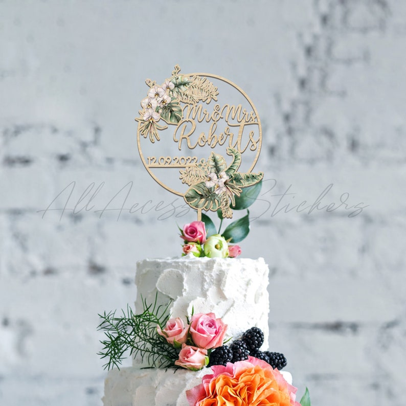 Peut inclure: Un cake topper en bois avec les mots "Mr & Mrs Roberts" et la date "12.09.2022" est entour&eacute; d'&eacute;l&eacute;ments floraux et de feuilles. Le topper est pos&eacute; sur un g&acirc;teau blanc &agrave; plusieurs niveaux d&eacute;cor&eacute; de roses roses, de verdure et de fleurs orange.