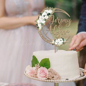 May include: A wooden cake topper shaped like a crescent moon, featuring the names Jeremy & Alyssa in script font. The topper is decorated with white floral accents and green leaves, and it is inserted into a white frosted cake on a silver stand.
