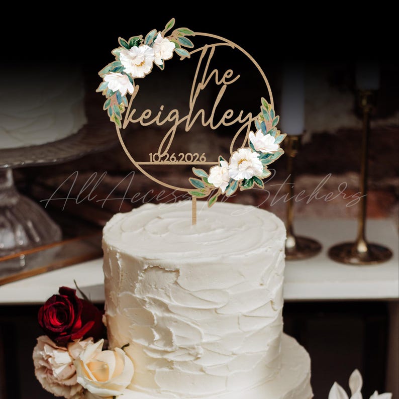 May include: A wooden cake topper with the words "The Keighley" and the date "10.26.2026" in a circular design. The topper is adorned with white flowers and green leaves. The topper is placed on top of a white frosted cake.