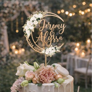 May include: A circular cake topper featuring the names Jeremy and Alyssa in a gold script font, with the date 07.26.023 engraved along the side, decorated with white floral arrangements on the top left and bottom right sections of the ring.